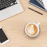 Cup of coffee with latte art on a wooden desk with laptop, phone, and pen.