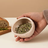 Hand holding a stone herb peeler bowl filled with green herbs on a neutral background