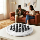 A round white marble game board with rows of black balls on a light wood table, with a blurred living room scene in the background showing a couple sitting on an orange couch.