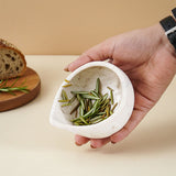 Hand holding a small cream bowl with green herbs on a beige surface.