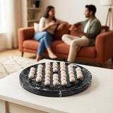 A round black marble game board with beige wooden pawns, shown on a white table in the foreground with a blurred living room scene behind it featuring two people sitting on an orange couch.
