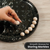 A hand places a smooth cream marble ball into a black marble tray with grooved recesses, with several matching marbles lined up on the side. The scene is set on a light wood floor, with a text overlay at the bottom right reading 'Smooth Groove for Storing Marbles'.