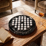 A round black marble boars with white marble veining, holding 36 small white spherical objects arranged in a grid, set on a rustic wooden coffee table in a living room with a glass of wine and an open book nearby.