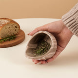 Hand holding a natural brown marble bowl with dried herbs, bread in the background