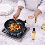 A chef in a white uniform seasons vegetables in a black frying pan on a gas stove, with a bowl of sliced potatoes, a bottle of oil, and stacked plates on a white marble countertop.