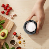 Hand reaching for a bowl of salt on a wooden table with ingredients like tomatoes, garlic, and avocados.