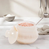 A cream-salt cellar with a matching lid, filled with pink Himalayan salt and a metal spoon, set on a white marble countertop with a neutral kitchen background.