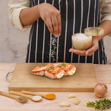 Person seasoning salmon on a wooden cutting board with various seasonings and herbs.