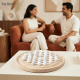A round tarvertine game  board with white balls  sits on a white table in the foreground, with a blurred couple sitting on a rust-colored couch in the background. The Earthen Crafts logo is visible in the top left corner.
