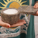Person holding a stone bowl with a lid filled with white powder against a colorful tapestry background.