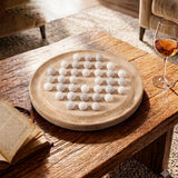 A round travertine stone board on a rustic wooden table, holding multiple small white polished stones. An open book, a wine glass with amber liquid, and part of a chair are visible in a warm indoor setting.