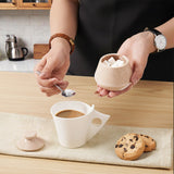 Person adding sugar to coffee in a kitchen setting with cookies on a wooden table.