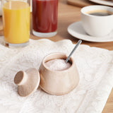 Small ceramic container of salt with a spoon on a white cloth, with drinks in the background.