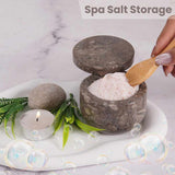 Spa salt storage with a wooden spoon, stones, and candles on a marble surface.