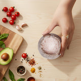 Hand holding a bowl of salad on a wooden table with ingredients like tomatoes, avocado, and spices.