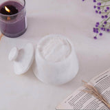 White marble container with lid on a light surface with a candle and flowers.