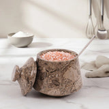 A decorative salt or pepper jar with a matching lid, filled with pink Himalayan salt and topped with a metal spoon, set on a light marble countertop in a kitchen scene.