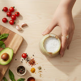 Hand holding a small bowl of salt on a wooden surface with tomatoes, garlic, and herbs.
