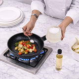 A chef in a white uniform adds salt from a glass bowl to vegetables cooking in a black frying pan on a gas stove, set on a white marble countertop with a bottle of oil nearby.