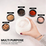 A hand holds a  salt cellar bowl filled with coarse salt, surrounded by small labeled bowls of ingredients including pepper, spices, pink salt, sugar, and almonds on a light surface. Text at the bottom reads 'MULTI PURPOSE' with descriptive copy.