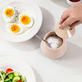 A marble salt container with a matching wooden lid is shown on a light tablecloth, with a hand lifting the lid. The container is filled with coarse white salt and a metal spoon is inside.