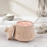 A beige stone-style salt or spice container with a matching lid, filled with pink crystals and topped with a metal spoon, set on a light marble surface in a softly lit kitchen scene.