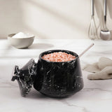 A black marble salt and pepper jar with its lid off, filled with pink Himalayan salt and a metal spoon, set on a white marble countertop with a neutral kitchen background.