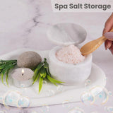 Spa salt storage with a bowl of salt, wooden spoon, candle, and leaves on a marble surface.