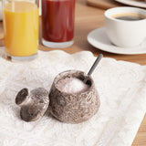 Decorative salt cellar with spoon on a lace tablecloth, with drinks in the background.
