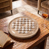 A round beige board holding a grid of small white marbled balls, styled on a rustic wooden table in a living room with a wine glass, book, and upholstered chairs visible.