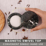 Hand holding a black marble container with a magnetic swivel top, surrounded by spices on a wooden surface.