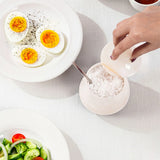 A hand is pouring coarse salt from a small white container with a spoon, set on a light textured tablecloth. Plates with food are partially visible around it.