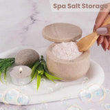  container of spa salt with a wooden spoon, candle, and leaves on a marble surface.