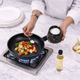 A chef in a white uniform seasons vegetables in a black frying pan on a gas stove, holding a black salt holder. A bottle of oil and other kitchen items are visible on a white marble countertop.