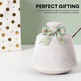 A white marble jar with a lid, decorated with a mint green floral ribbon bow. A gold spoon rests inside, and the background features a sage green wall, a white gift bag with gold polka dots, and promotional text reading 'PERFECT GIFTING'.