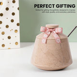 A beige  jar with a lid and a pink ribbon bow with white flowers, set on a white surface with a green background and text reading 'PERFECT GIFTING'.