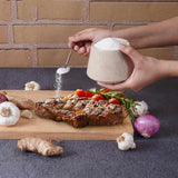 Person seasoning a steak with salt on a wooden board, surrounded by vegetables and a brick wall.