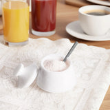 A small white sugar bowl with a lid and spoon, filled with white sugar, set on a white lace placemat with breakfast items in the background.