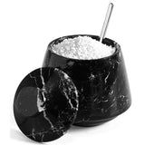 A black marble salt and pepper container with a matching lid leaning against it, filled with white salt, and a metal spoon resting inside, on a white background.