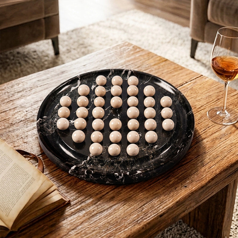 A round black board game  styled on a wooden table in a living room setting with a wine glass and an open book nearby.