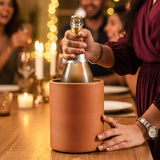 Person opening a bottle of champagne with a decorative cork in a terracotta cooler, surrounded by people and lights.